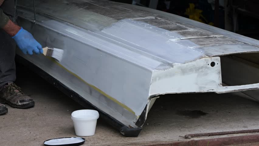 A person paints the underside of a small fiberglass boat with a brush, showing surface preparation and careful coating during a DIY marine restoration process.
