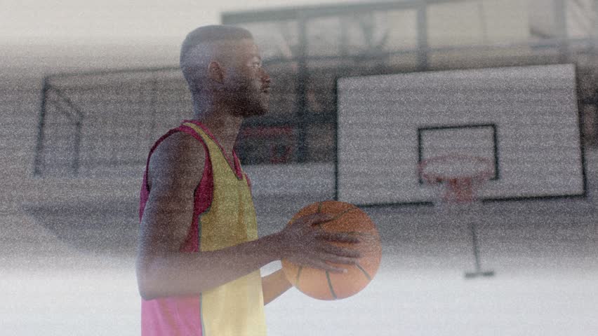 Man holding basketball shifting, aiming at hoop and turning to camera, overlay drifting across man. Court, ball, athlete, jersey, backboard, rim, grainy