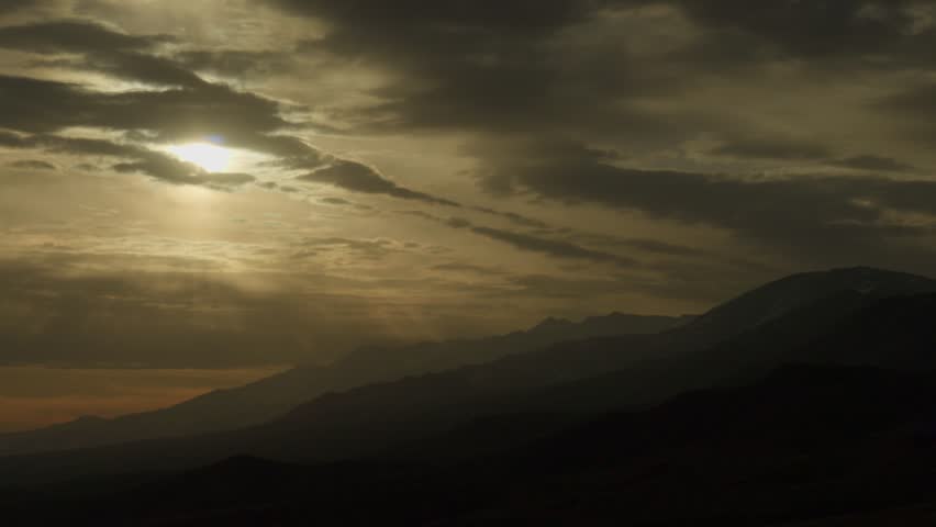 Dramatic sunset over mountain range with strong sun rays breaking through clouds. Atmospheric landscape with silhouettes of hills and layered mountains, moody sky and warm golden light.