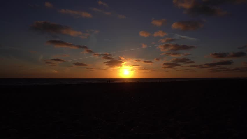 Stunning wide-angle footage of a golden sunset over the North Sea from a sandy beach near Billund, Jutland, Denmark. The sun sets directly on the horizon, casting a brilliant orange and golden glow across the sky with scattered lit clouds. Silhouetted figures of people are visible near the water