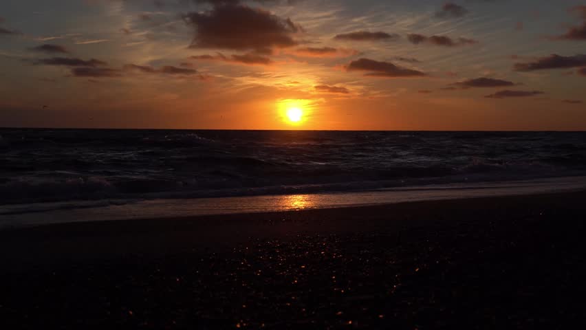 Dramatic footage of an intense orange sunset over the North Sea near Billund, Jutland, Denmark. The bright orange sun glows just above the horizon while dark waves roll onto the wet sandy beach, reflecting the golden light. Scattered birds fly across the orange sky, and clouds are lit in deep amber and red, creating a powerful and atmospheric evening scene.