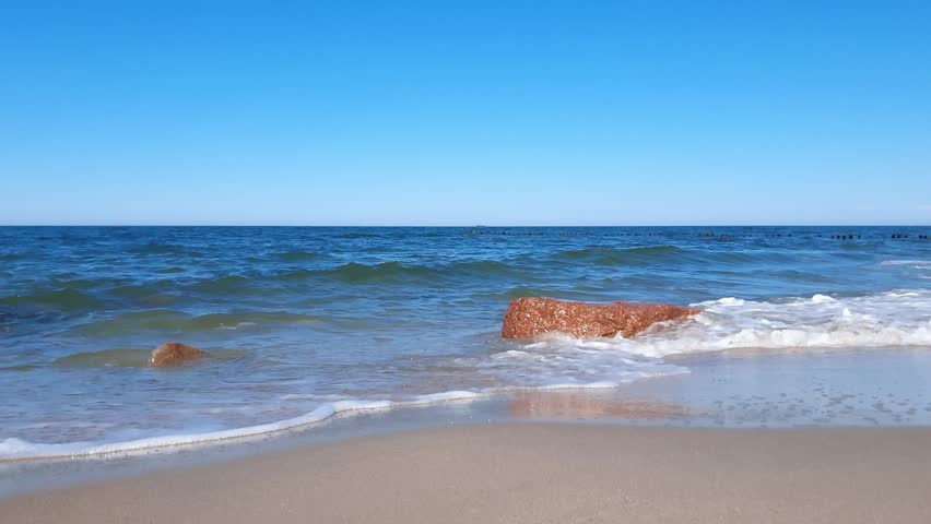 Tranquil seaside landscape of Baltic sea shore with calm turquoise coastal surf water waves rolling on boulder and empty sandy beach under clear blue sky at sunny day. No people seascape background.
