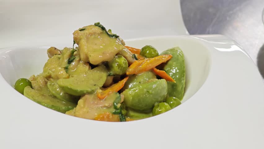 Close-up of pouring rich Thai green curry sauce over chicken and eggplant in a white bowl.
