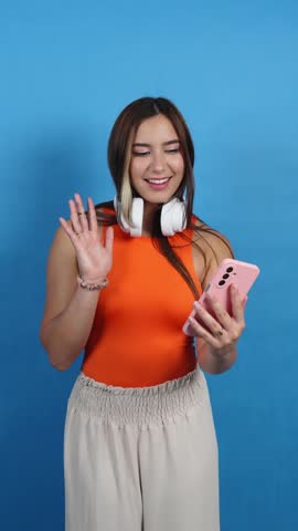 Cheerful woman with headphones around her neck smiling and waving at her smartphone on a blue background