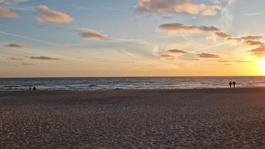 Ground-level panoramic footage of a wide sandy beach on the North Sea coast near Billund, Jutland, Denmark. The sun sets over the ocean with warm golden and orange hues illuminating the clouds. Silhouettes of visitors stand near the water while contrails cross the expansive evening sky. Shot in summer on August 1, 2022.