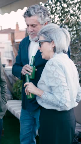 Group of smiling mature people enjoying a conversation while drinking beer. Elegant friends gathering on a terrace for a social event. Vertical