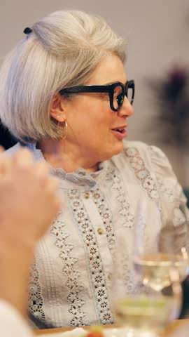 Stylish mature woman with grey hair and glasses enthusiastically talking and gesturing at a dinner table. She enjoys a lively conversation. Vertical