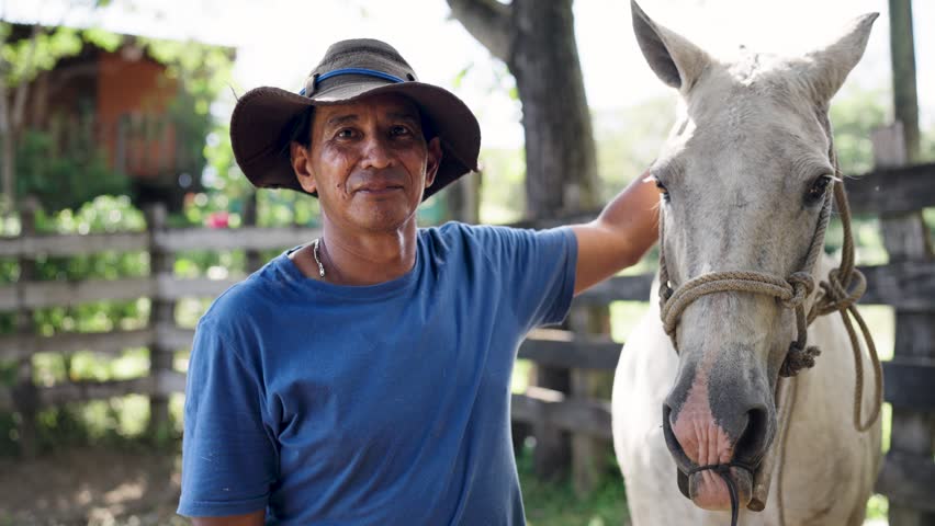 Hispanic man with a hat smiling and caressing his white horse. Rancher showing his affection for his animal on a farm in Latin America