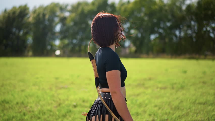 female archer walking across sunlit meadow, side profile of contemplative woman wearing black crop top and studded leather fringe skirt with quiver on back, warm golden light, soft breeze ruffling