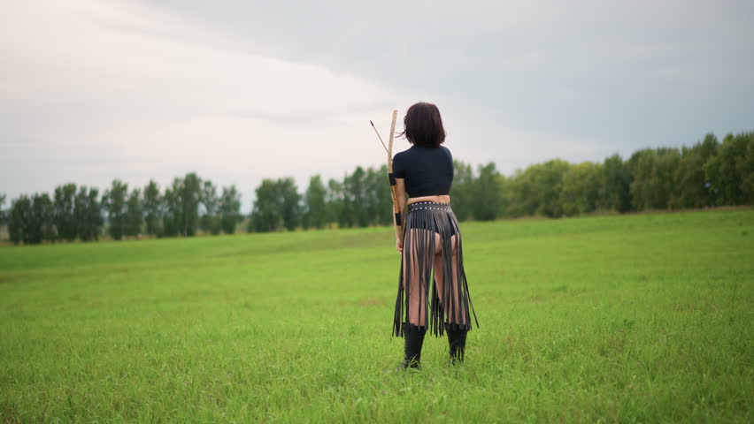 white woman warrior stands in green field, leather skirt and bow on back, facing tree line under overcast sky, contemplative stance suggesting solitude and readiness, scout scanning horizon, ranger