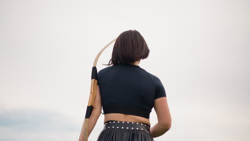 female archer outdoors drawing bow, steady stance and focused aim under cloudy sky, leather quiver at hip, recurve bow pulled back with arrow nocked, tense moment before release conveying calm
