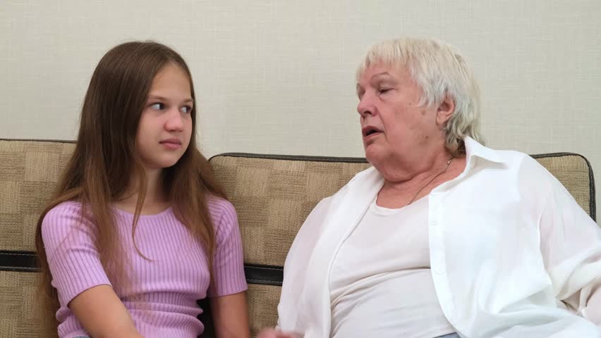 Senior woman scolding and gesturing while teenage girl listens attentively on couch at home. Grandmother scolds her granddaughter indoors. Concept of family communication