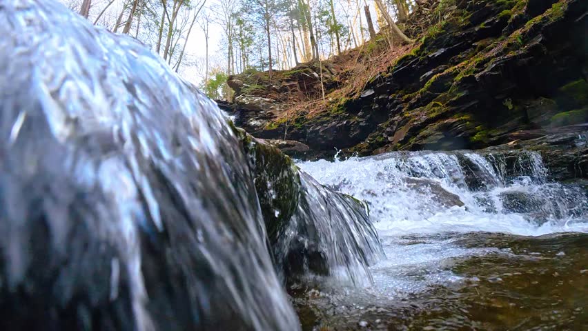 Cascading waterfall over layered dark rocks in forest stream. Close up of water flowing over tiered shale rock formations in a woodland environment.