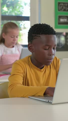 Vertical video: Typing boy in yellow top slowing resting at classroom desk from fatigue with laptop. Student, study, computers, daylight, school, focus, vertical video