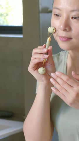 Vertical video: Picking jade roller, Asian woman rolling cheek and under-eye at vanity, green top. Facial, skincare, closeup, massage, bathroom, mirror, vertical video