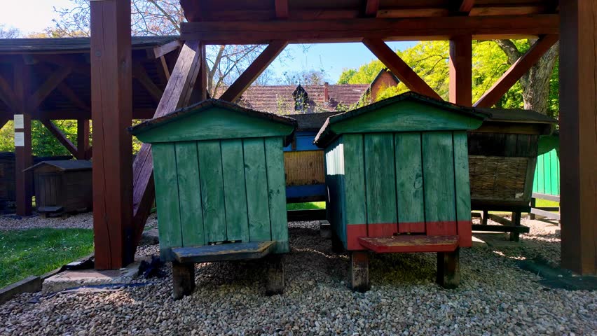 Vintage wooden beehives painted in green and blue with red accents, set on gravel under a protective wooden shelter in an open-air museum, with old tiled building roofs and spring green trees visible