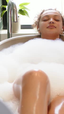 Vertical video: Camera starting low filming woman soaking, relaxing in tub by window, foam, earbud. Bathroom, bathtub, bubbles, serenity, backlit, greenery, vertical video