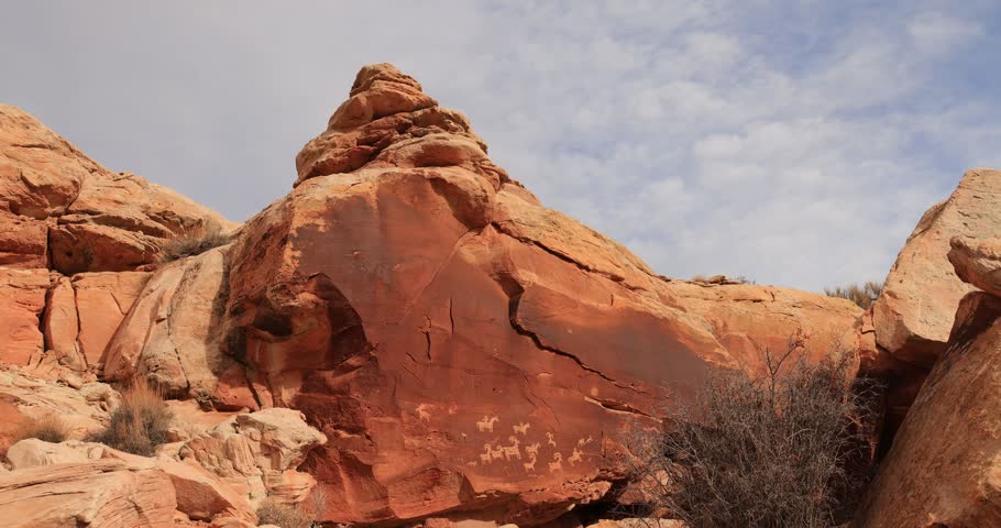 Large reddish-orange rocks with stylized animal carvings sit in a sunlit desert environment.
