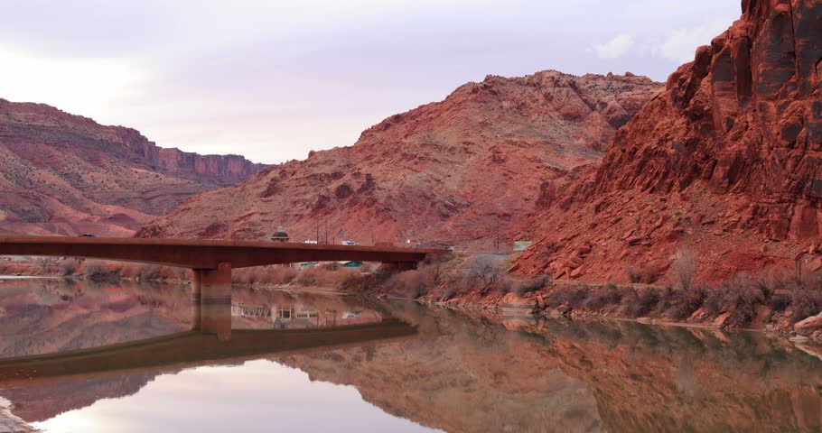 Water flows slowly beneath a bridge connecting steep, red canyon walls under a soft sky.
