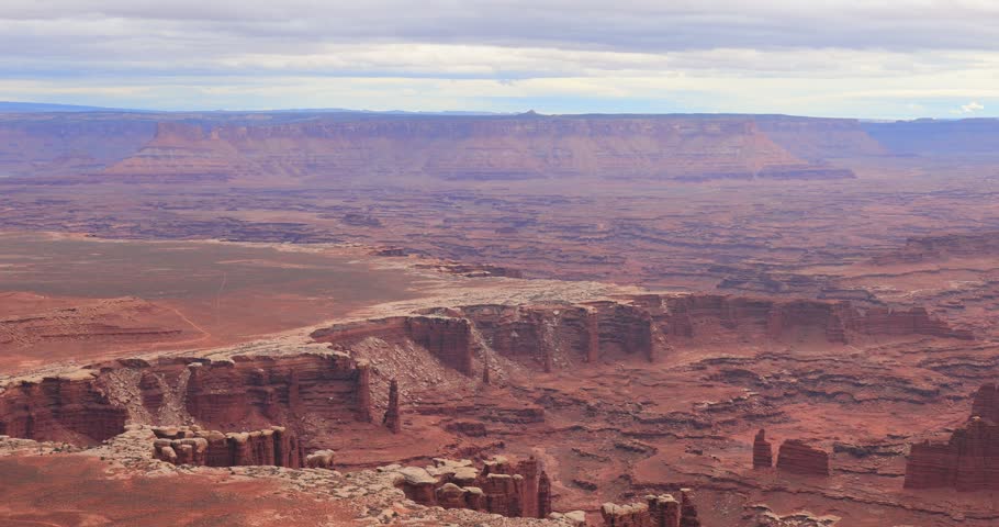 A sweeping panoramic view captures the vast red rock desert landscape and towering cliffs of Arches National Park under a cloudy sky.
