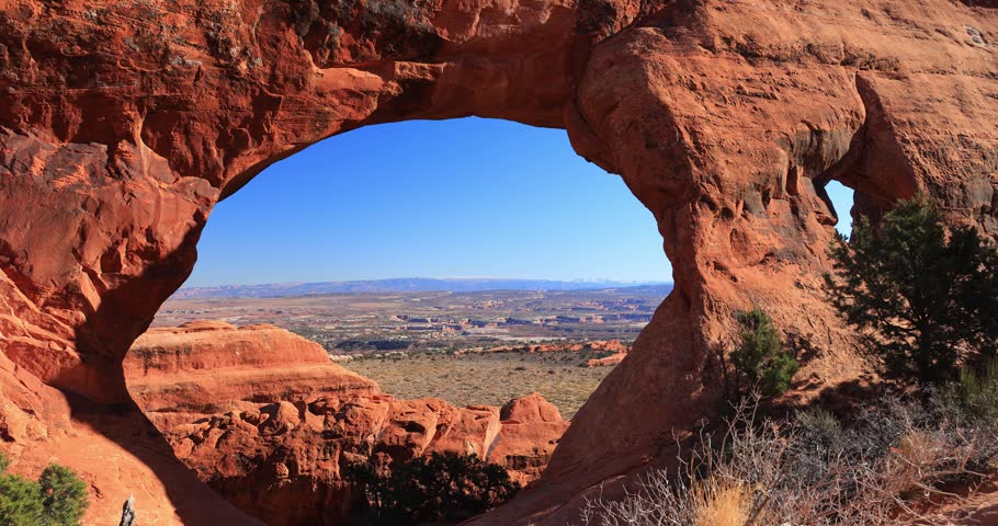 A large natural red rock arch frames a sweeping view of a rugged canyon and distant mountains under a clear blue sky.