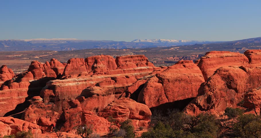 Small figures walk among towering red sandstone cliffs under a clear blue sky with distant snow-capped mountains visible on the horizon.