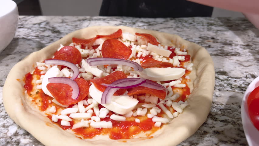 The video shows hands placing onion slices across the top of a sauced and cheesed pizza dough. Soft indoor lighting highlights the food preparation process on a granite countertop.