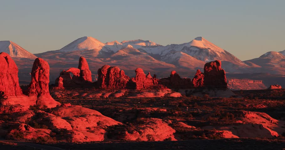Sunlight bathes towering red sandstone arches in a deep crimson glow while distant snow-capped La Sal Mountains rest under a clear evening sky.