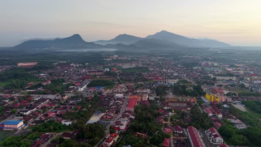 Aerial view of quiet town at dawn. Red roofs glow under soft sunrise light. Mountains loom softly in the misty background at Tangkak