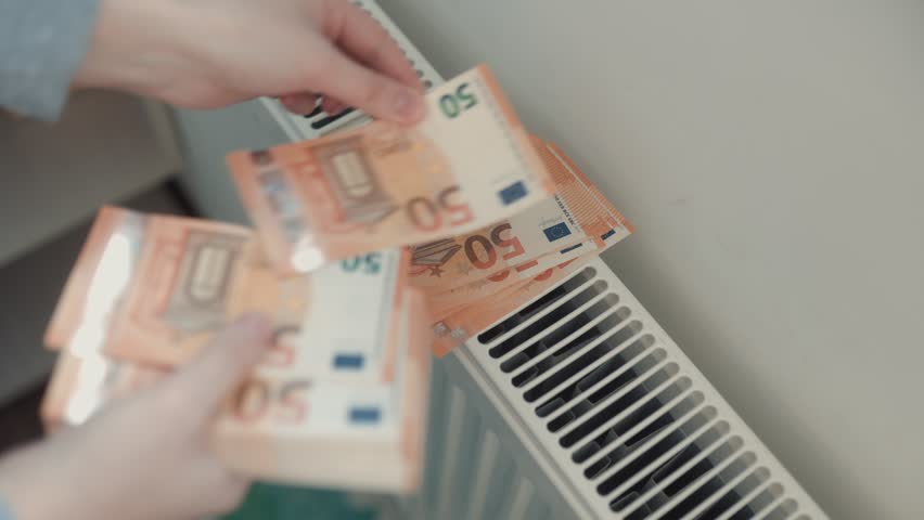 Close up shot of hands placing several fifty euro banknotes on a white domestic radiator, illustrating the high cost of energy, heating bills, and inflation crisis.