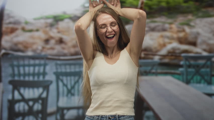 Woman making rock horns with hands near face, smiling with glasses and sleeveless top on a cafe terrace at a building; playful rebellion.