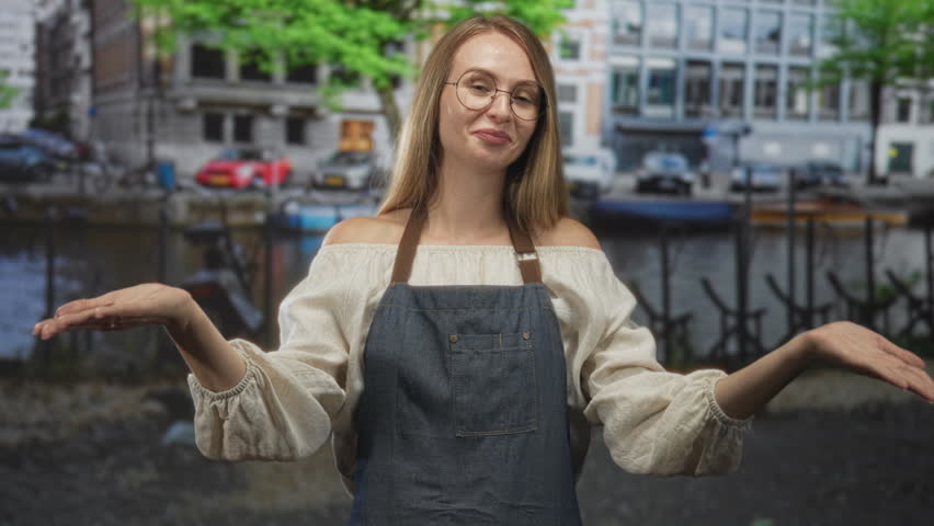 Woman in apron offers palms up and smiles while gesturing toward camera on a city street by a canal; welcoming.