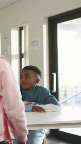 Vertical video: Passing girl in pink hoodie, braid swaying left at school, boy in blue watching. Teens, peers, classroom, campus, hallway, posters, vertical video
