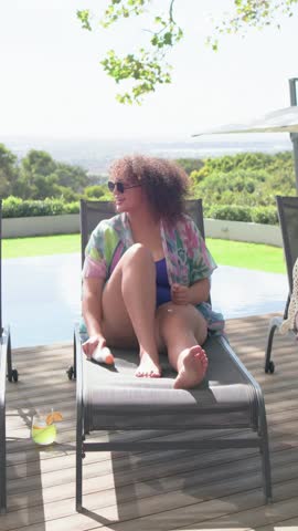 Vertical video: Reaching woman in blue swimsuit applying sunscreen to legs at poolside for sun care. Pool, deck, outdoors, vacation, wellness, skincare, vertical video