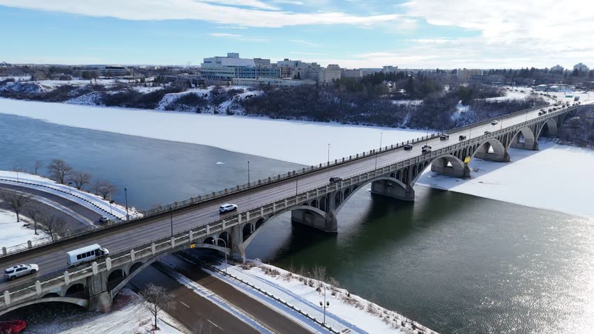 Aerial drone captures a snowy bridge over a partially frozen river. Vehicles travel across the arches in winter daylight. Snow blankets nearby city buildings and trees