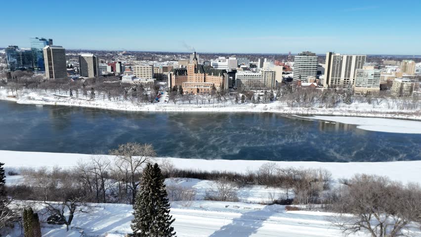 Clear blue sky shows downtown Saskatoon in winter. Frozen river flows beneath snow-covered banks. City buildings stand against bright horizon. Drone captures quiet urban landscape