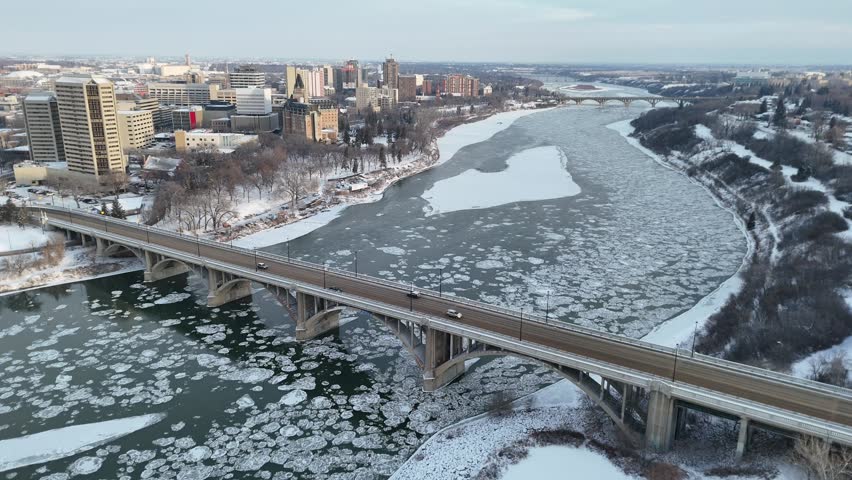 Aerial view captures downtown Saskatoon. Bridge spans a frozen river. Ice floes float on the water. Snow blankets city streets. Cars travel across the bridge. Winter urban landscape with clear skies