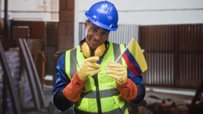 Man construction worker in hardhat and hi vis holding colombian flag, finger to lips in building; pride support safety.
