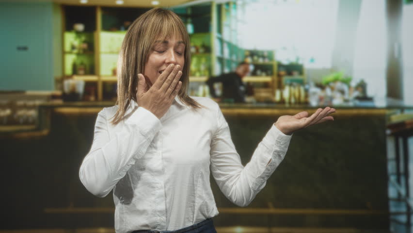 Woman covers mouth with hand and displays palm up gesture by a bar counter in a building; surprise delight.