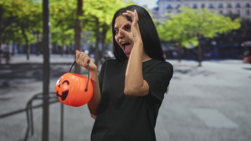 Hispanic woman holding orange pumpkin bucket on street, hand extended and making ok circle with fingers near eye while smiling; festive playful joy.
