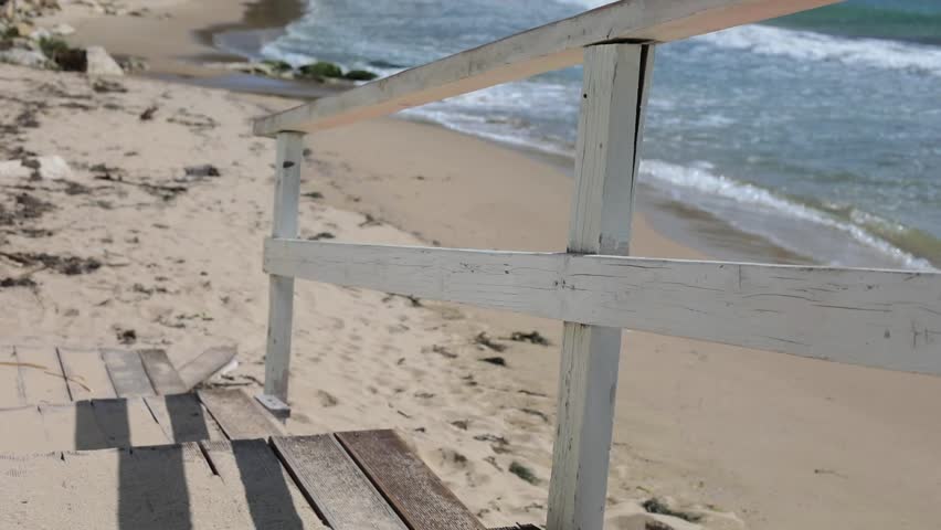 old weathered white wooden railing and steps leading down to sandy beach with sea waves breaking on shore and footprints in sand