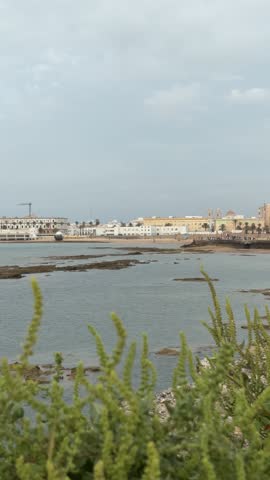 Cadiz, Andalusia. Panoramic view of a coastal city featuring traditional architecture along a waterfront promenade. The scene shows calm sea waters, low-rise buildings in warm tones, and a peaceful seaside atmosphere.