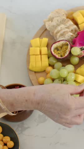 Vertical video: Hands entering, host in rust top arranging wood tray on counter, slicing grapefruit. Fruitplatter, mango, dragonfruit, passionfruit, grapes, pomegranate, vertical video
