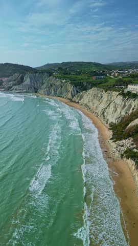 Coastal landscape of Sicily featuring sandy beach, turquoise waves, and steep cliffs with greenery and homes visible along the shoreline