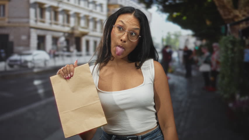 Young latina woman holding a brown paper bag and sticking her tongue out on a busy urban street, wearing glasses and a white tank top; playful.