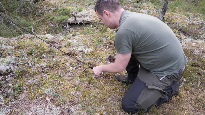 Outdoor scene of a man kneeling on moss and lichen in a woodland area, handling a cord or line during hiking, camping, survival, or nature activity.