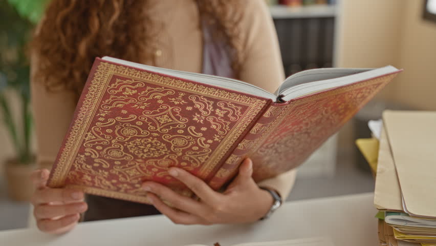 Woman reading an ornate red book, wearing glasses and hoop earring, finger to lips for silence in building; contemplative hush.