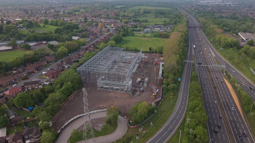 Aerial drone forward flight approaching a commercial building under construction near a motorway. Industrial development, real estate and infrastructure growth concept.