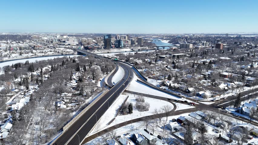 Aerial view of Nutana Street in Saskatoon. Snow blankets residential areas and city skyline. Curved highway flows through the scene. Clear winter sky shows active traffic movement