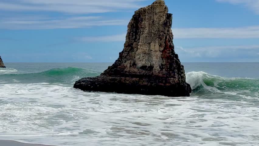 Powerful ocean waves crash against a rugged sea stack along the Pacific Coast. Scenic coastal landscape with textured rock formation, blue sky and dynamic surf capturing beauty of California shoreline